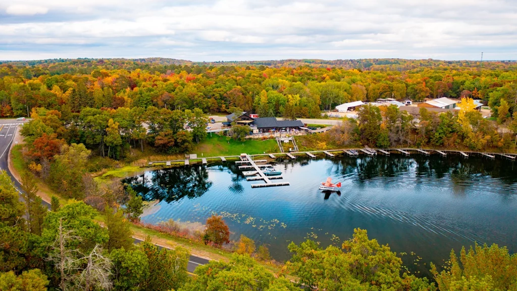 Photo of a lake on a fall day surrounded by forest with docks at the edge of the water and a plane landing on the lake with a restaurant on the hill above the water