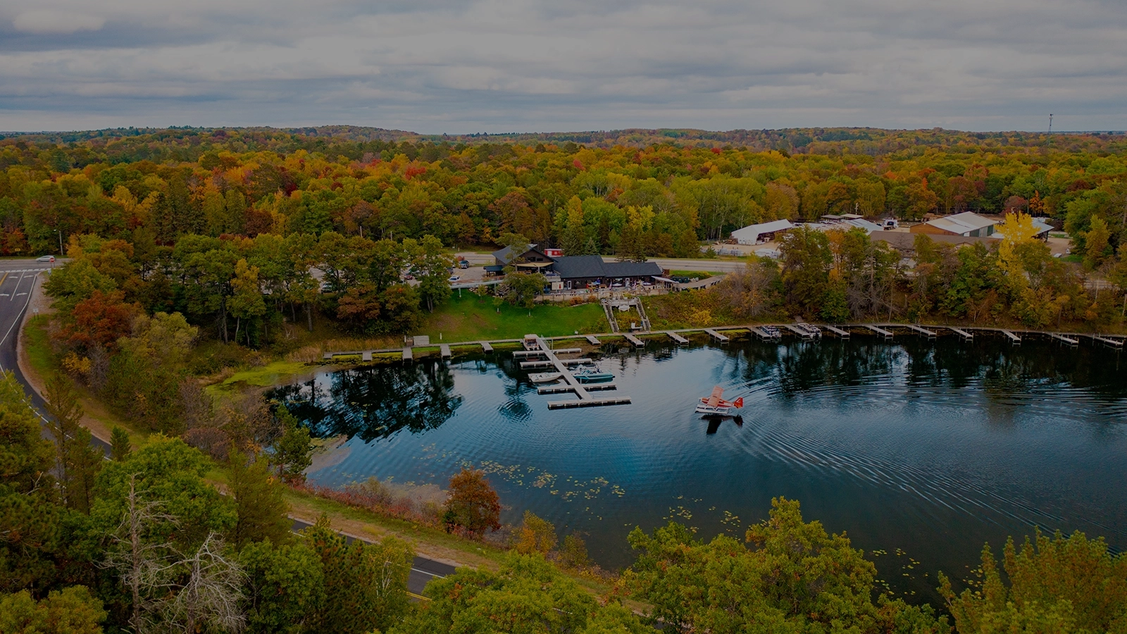 Photo-of-a-lake-on-a-fall-day-surrounded-by-forest-with-docks-at-the-edge-of-the-water-and-a-plane-landing-on-the-lake-dark.webp