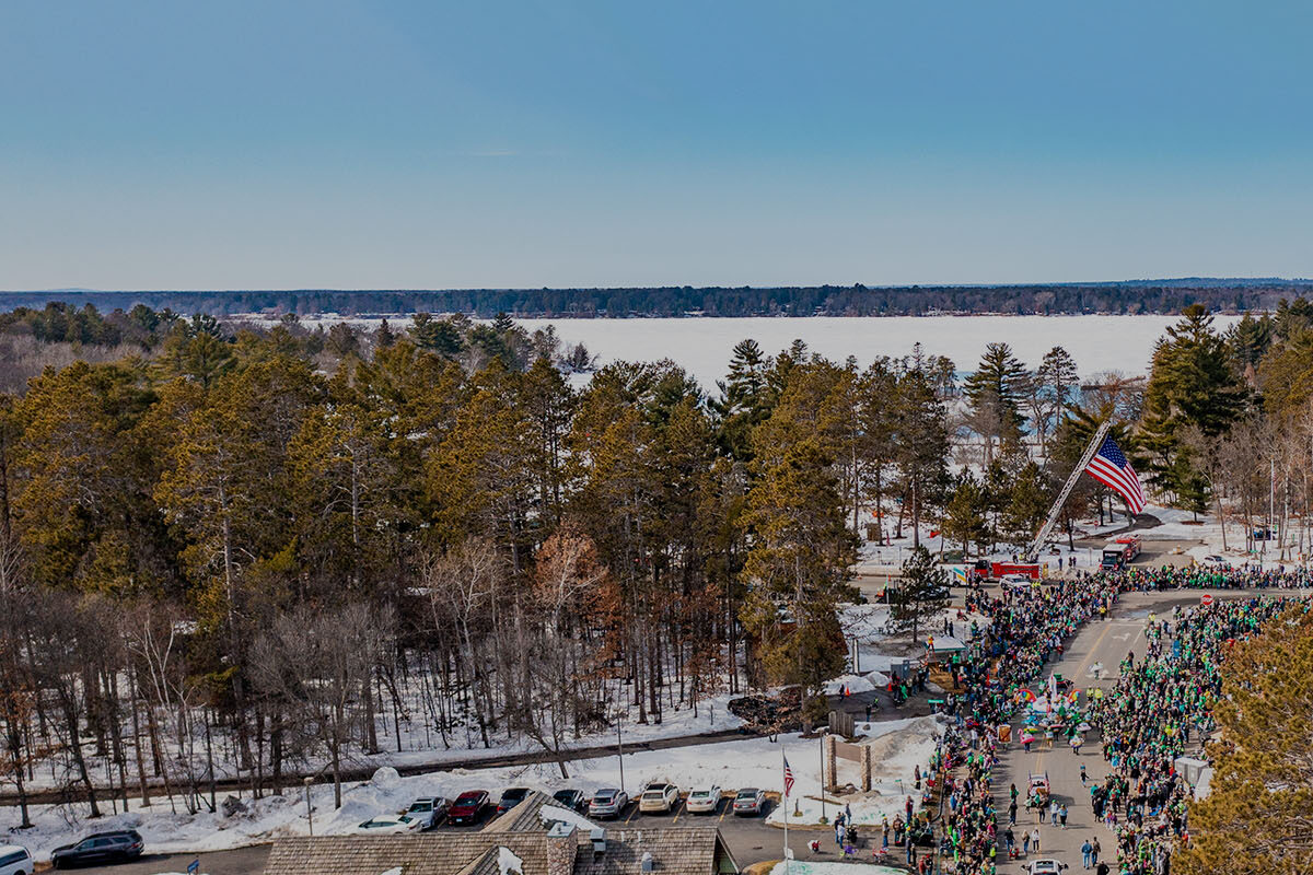 This image shows the town of Crosslake, MN on a winter day during the St. Patrick's Parade. It shows an aerial view of the crowds down a street with trees around and a lake in the distance. There is a flag flowing in the middle of the image above the street.